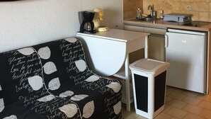 A compact kitchen with a white countertop, a sink, and a refrigerator. A black sofa with white leaf patterns is positioned against the wall.
