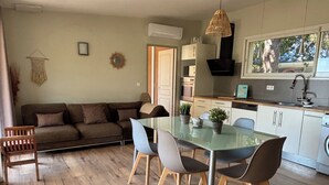Un salon moderne avec une table à manger en verre, des chaises en bois, un canapé marron et un coin cuisine doté de meubles blancs et d’un luminaire suspendu.