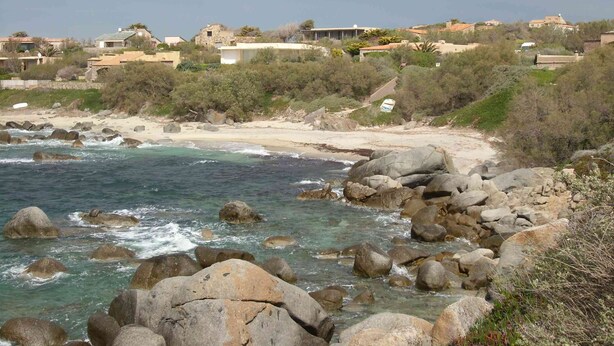 Een kustlandschap met rotsachtig terrein, een zandstrand en gebouwen op de achtergrond.