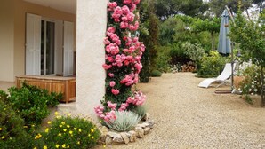 Een tuin met een grindpad, een houten bankje en een rij roze bloemen tegen een gevel.