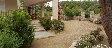 Uma casa moderna com varanda coberta, entrada com calçamento de pedras e jardim com diversas plantas e flores.