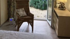 Een slaapkamer met een rieten stoel, een bed, een nachtkastje met een lamp en uitzicht op een tuin door een schuifdeur.