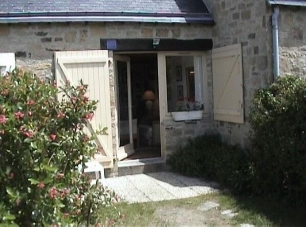 A stone building with a wooden door and window shutters, a small porch, and a garden area with bushes and a flowering plant.