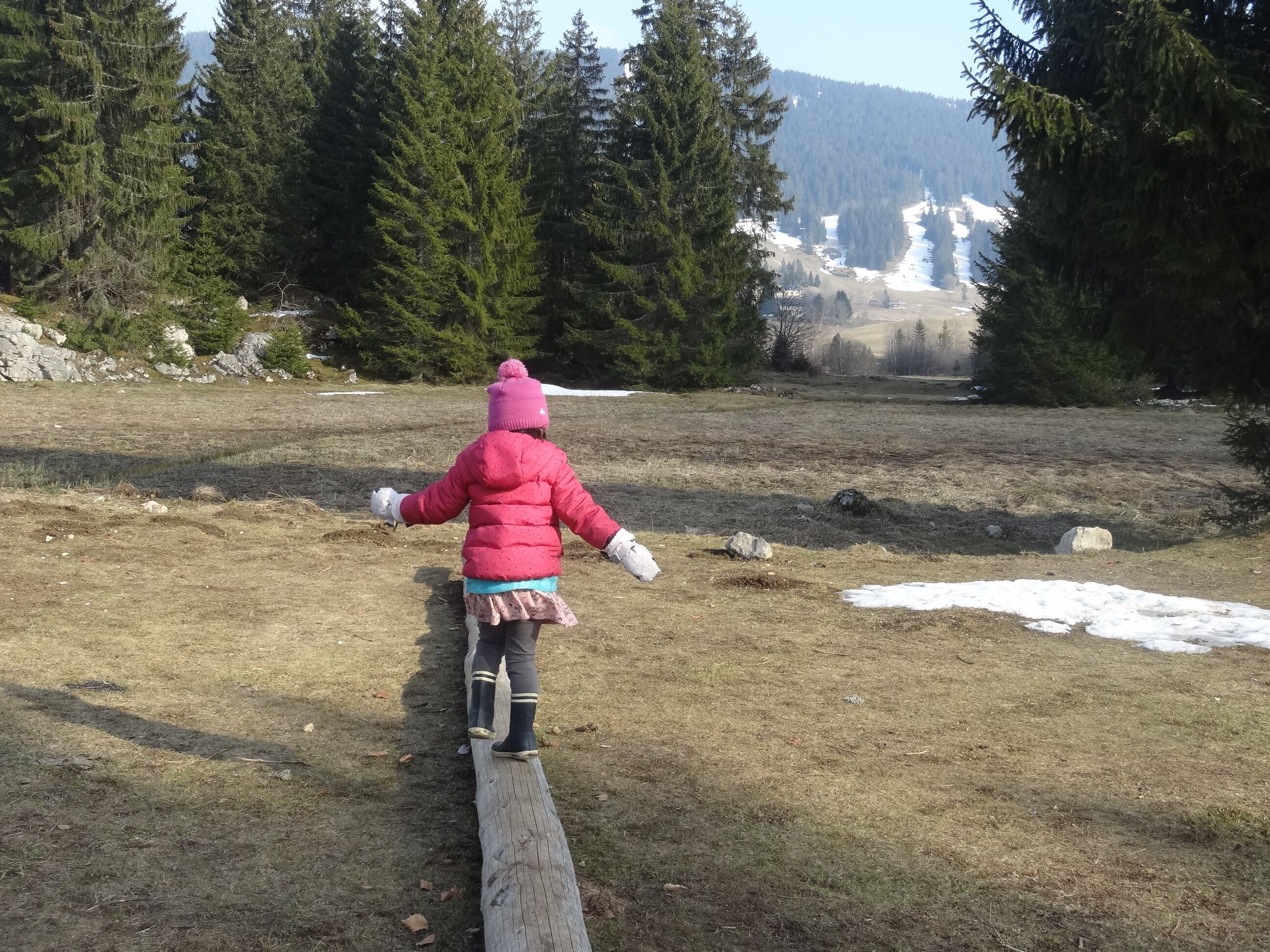 Ein Kind geht auf einer Holzbrücke in einem bewaldeten Gebiet mit Schneeflecken und Bäumen.