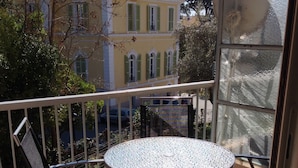 En balkon med et rundt bord, en stol, gelænder og udsigt til en gul bygning med grønne skodder.