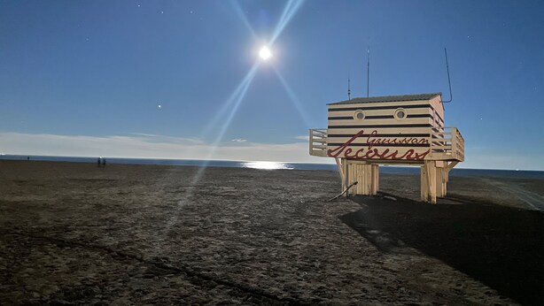 Torre de socorrista con las palabras 'Lifeguard' y 'Secours', ubicada en una playa con cielo despejado y el sol visible.
