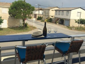A balcony with a table set for two, featuring a bottle and two glasses. There are wicker chairs and a hat on the table.