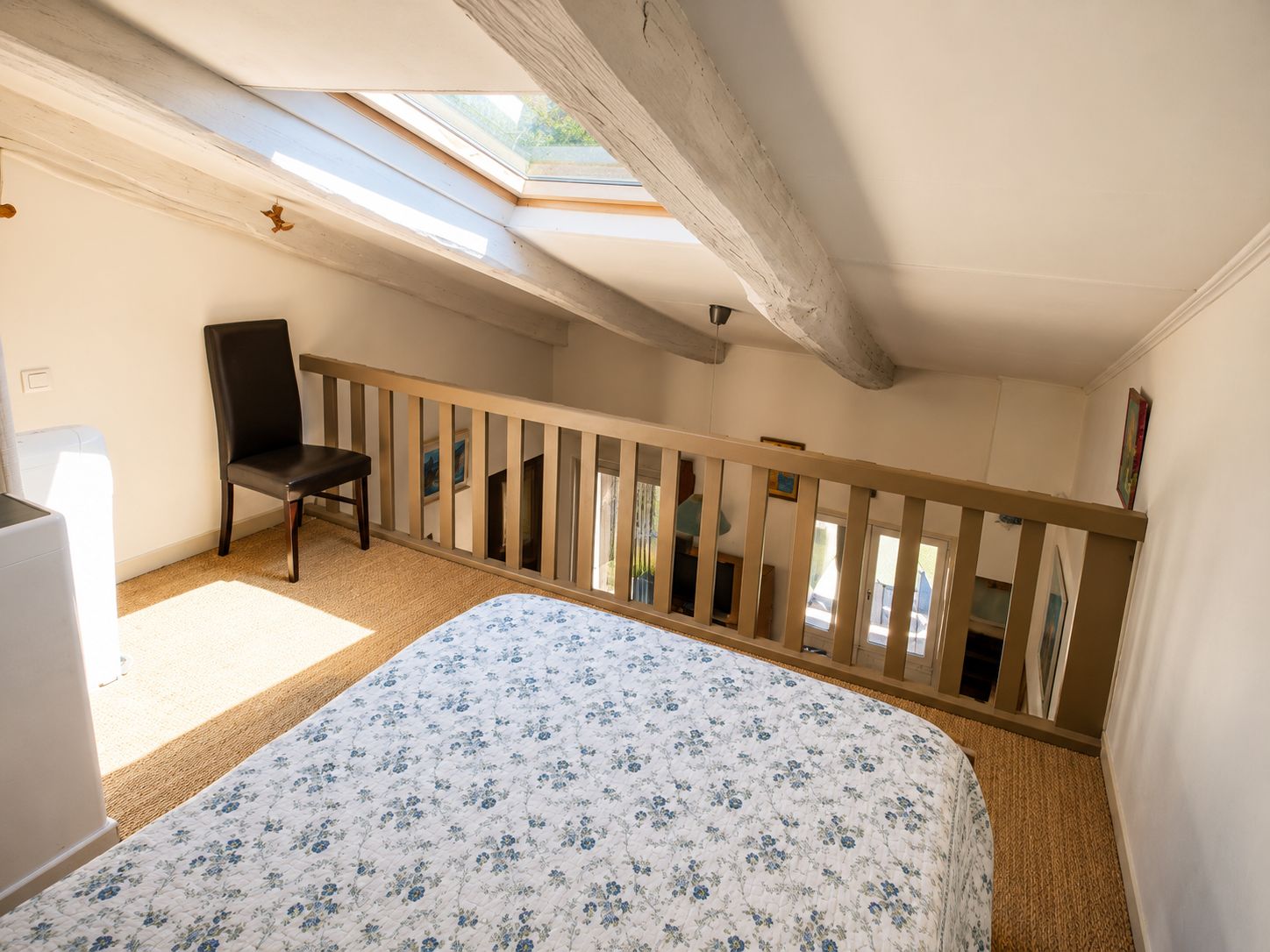 A bedroom with a skylight, a bed with a floral duvet, a chair, and a wooden railing.