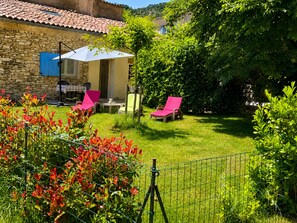 A stone building with a tiled roof, a covered patio with outdoor furniture, and a lush garden with a fence.