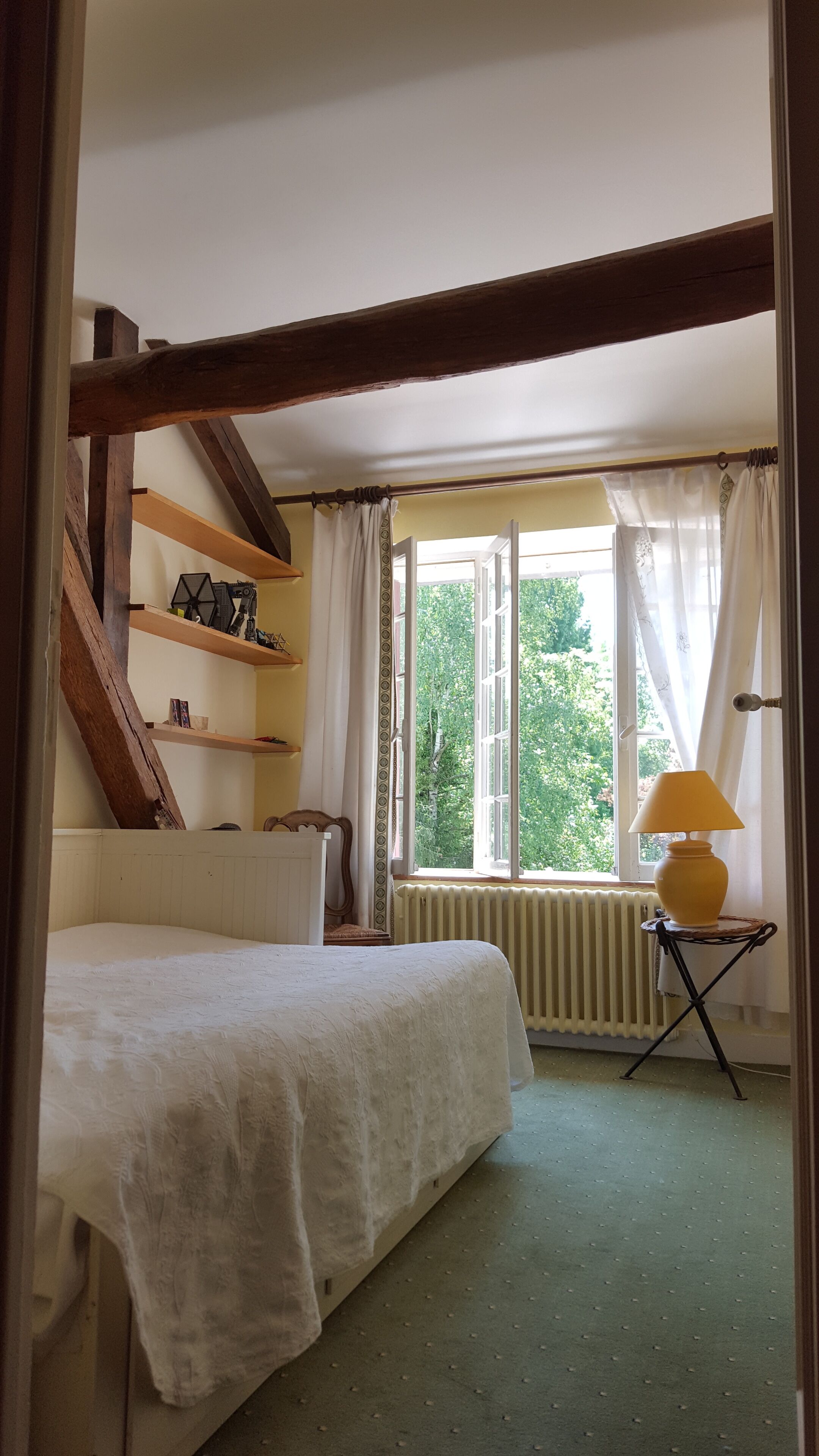 Un dormitorio con cama, ventana con cortinas, radiador y lámpara sobre una mesita.