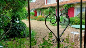 A garden with a gravel path, a metal gate, and a building with red shutters and a tiled roof.