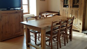 Une table à manger en bois, accompagnée de six chaises, un téléviseur posé sur un meuble en bois, et une fenêtre avec des stores.
