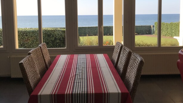 A dining area with a table set for four, large windows offering a view of the sea, and a hanging red light fixture. - Grandcamp-maisy sea view apartment, direct beach access, 4 rooms, sleeps 6 (Grandcamp-Maisy)
