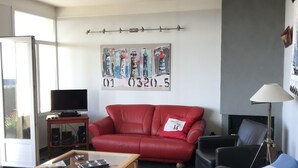 A living room with a red sofa, black armchair, and a coffee table. There is a television on a stand, a painting on the wall, and a lamp. - Grandcamp-maisy sea view apartment, direct beach access, 4 rooms, sleeps 6 (Grandcamp-Maisy)