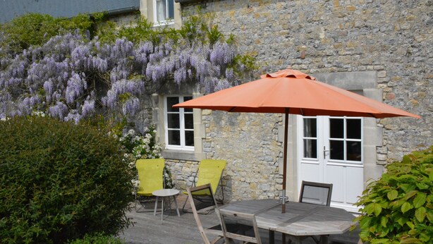 Un bâtiment en pierre doté d’une terrasse en bois, d’une table et de deux chaises jaunes.