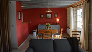 Un coin repas avec une table ronde en bois, des chaises et un vase de fleurs. On aperçoit un canapé au premier plan et une fenêtre avec des rideaux sur la droite.