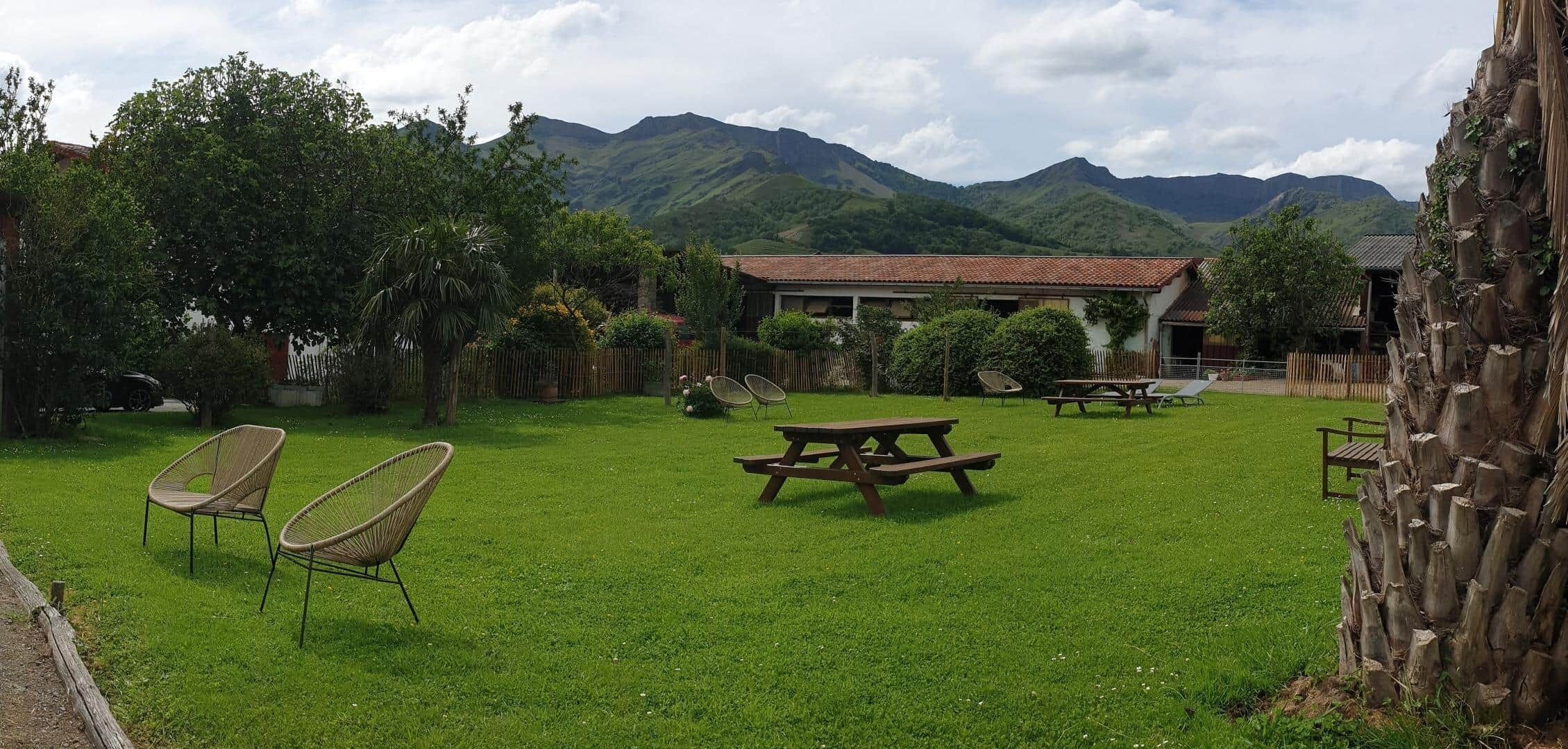 A grassy outdoor area with picnic tables and chairs, surrounded by trees and a building with a red roof.