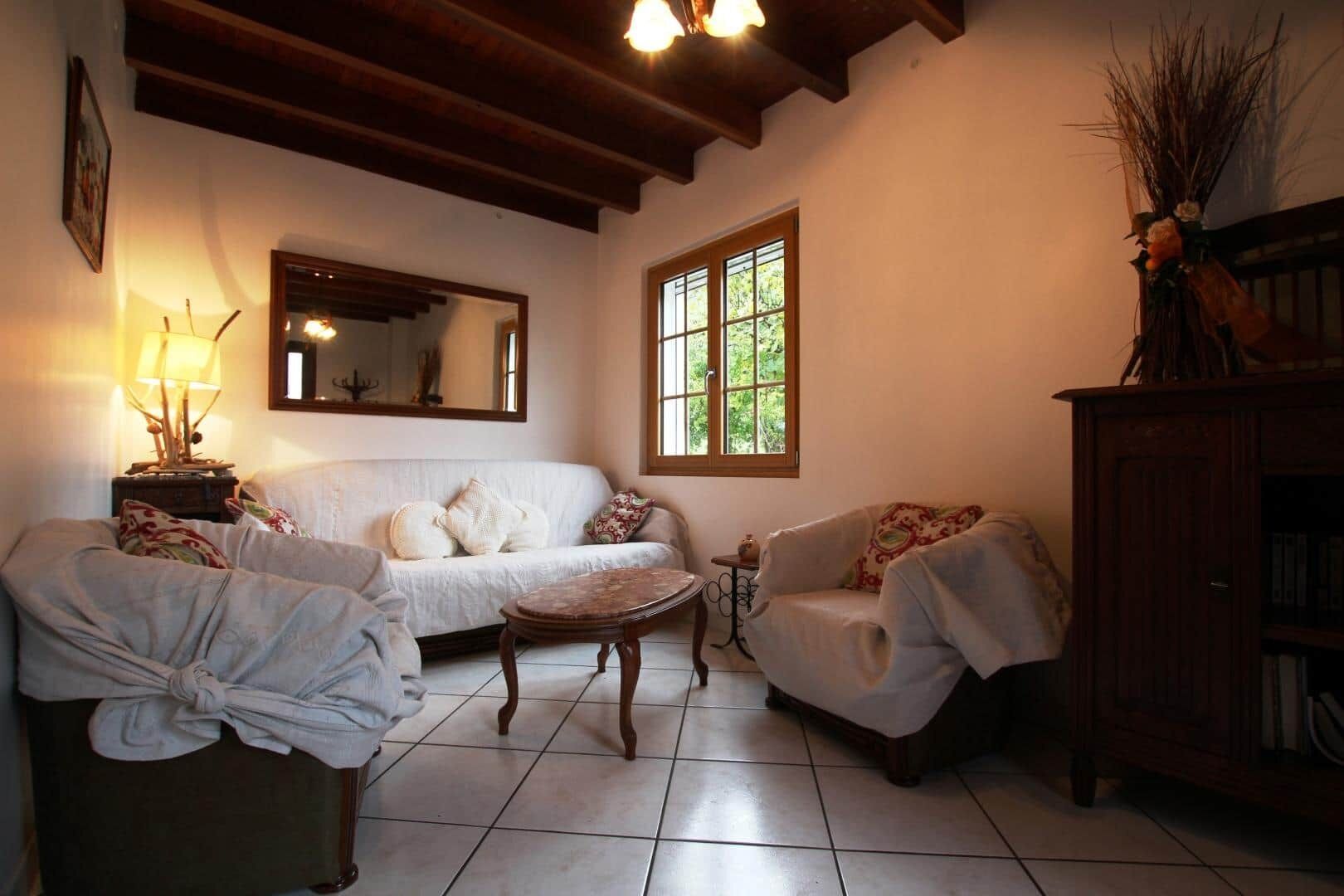 Una sala de estar acogedora con un sofá blanco, una mesa de centro de madera y una silla. Se observa un aparador de madera con libros y un espejo en la pared.