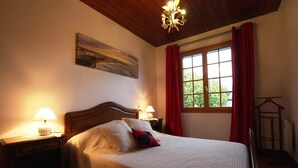 A bedroom with a large bed, two bedside tables with lamps, a painting on the wall, a window with red curtains, and a wooden ceiling.