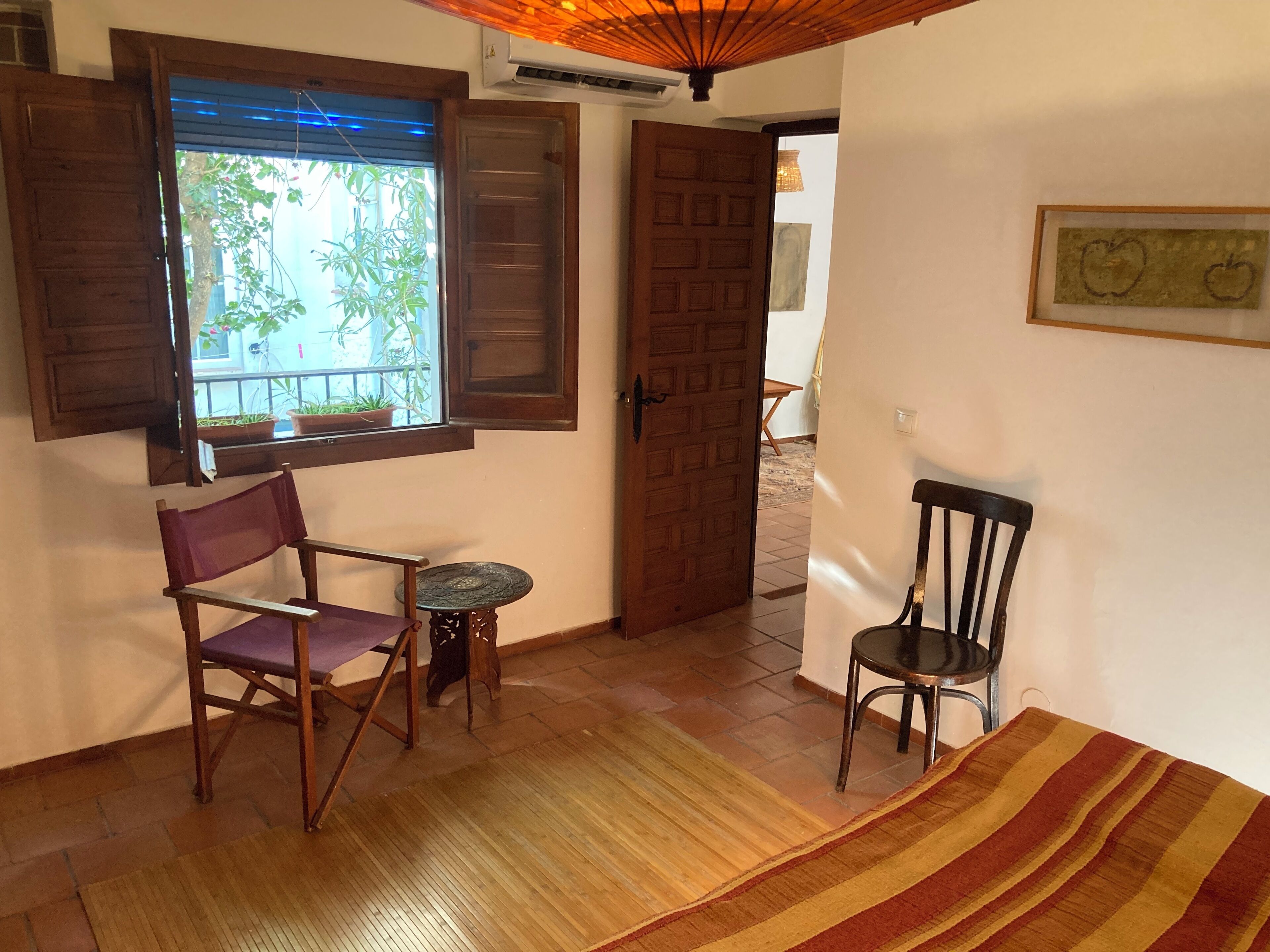Una habitación con piso de madera, una pequeña mesa redonda, una silla morada, una puerta de madera y una ventana con contraventanas de madera.