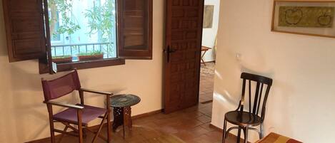 Una habitación con piso de madera, una pequeña mesa redonda, una silla morada, una puerta de madera y una ventana con contraventanas de madera.