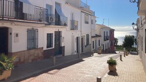A narrow cobblestone street lined with traditional buildings, some with balconies and satellite dishes, leading towards a body of water. - Village house with large rooftop terrace near the beach (Maro - Nerja)