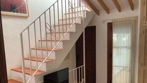 A staircase with white railings and wooden steps, a flat-screen TV mounted below, a door with a wooden frame, and a window with a sheer curtain. - Village house with large rooftop terrace near the beach (Maro - Nerja)