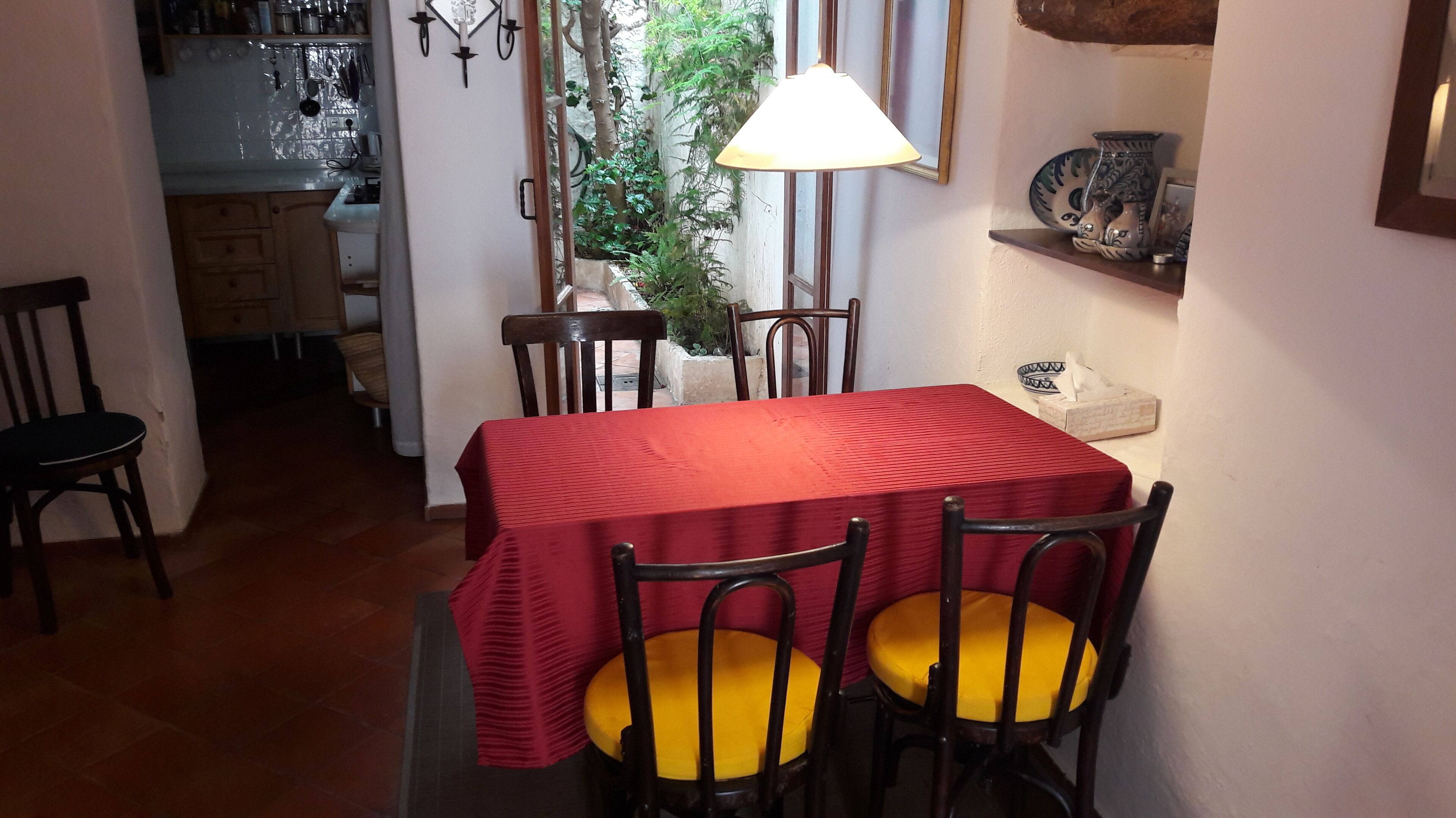 A dining area with a red table and yellow cushioned chairs, a hanging lamp, and a shelf with decorative items.