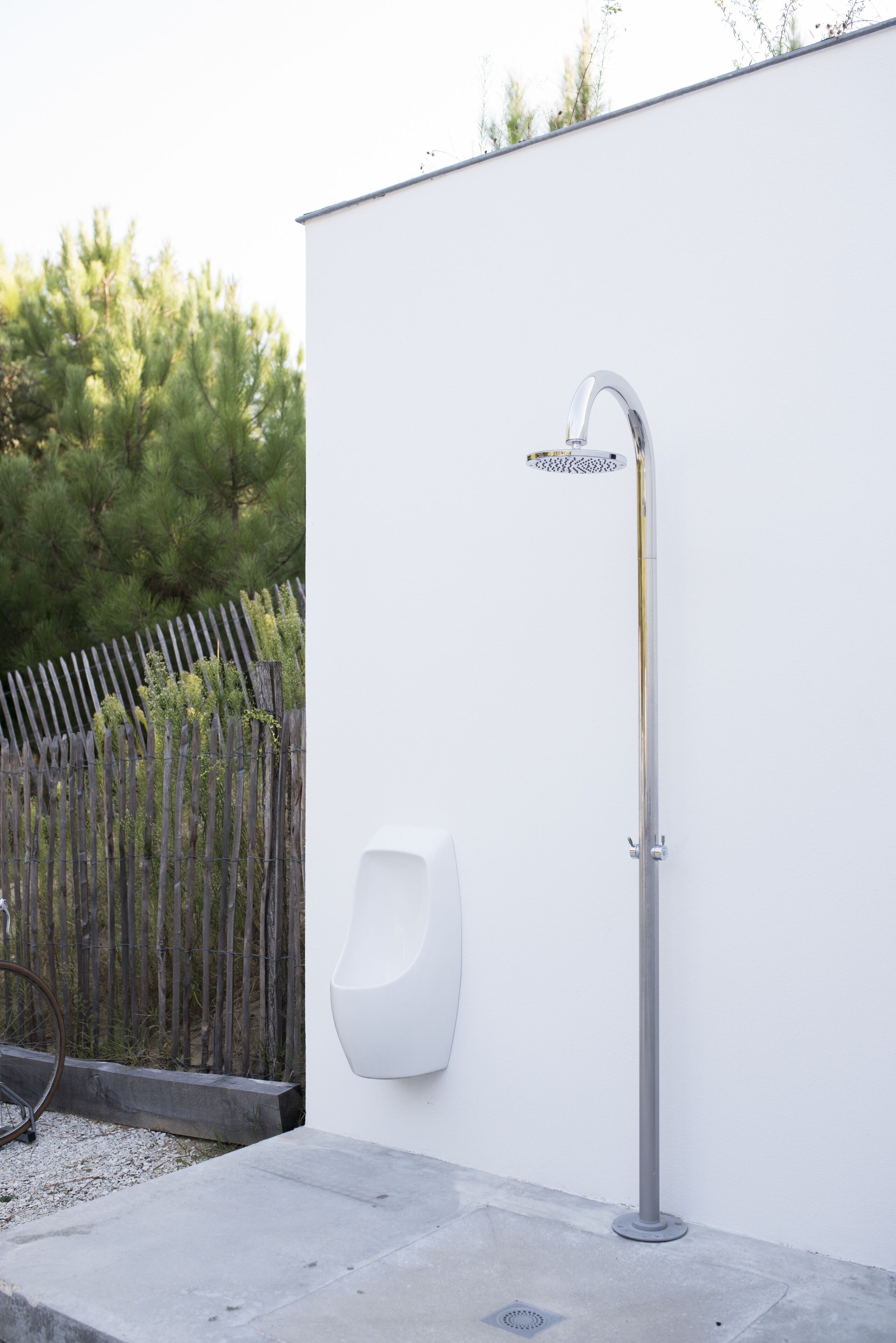 A modern outdoor shower with a high-arc faucet, a wall-mounted urinal, and a wooden fence in the background.