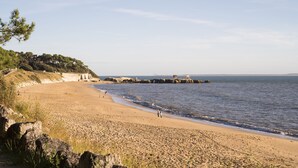 A beach with a rocky shoreline, a few people walking, and a distant view of a cityscape. - THE DUNE VILLA PLATIN ST PALAIS SUR MER (Saint-Palais-Sur-Mer)