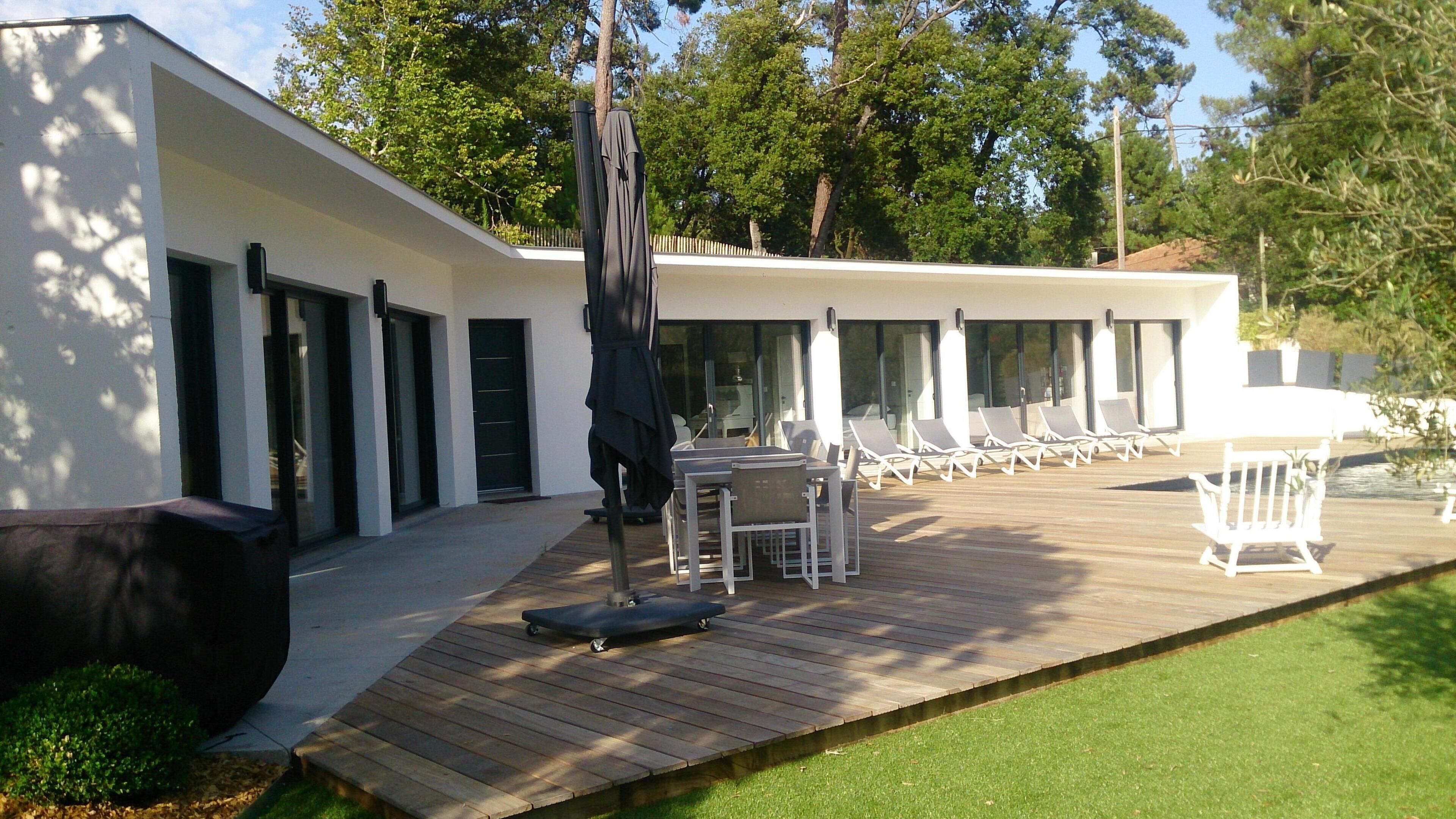 A modern outdoor patio with a wooden deck, a dining table with chairs, and a lounge area with sun loungers.