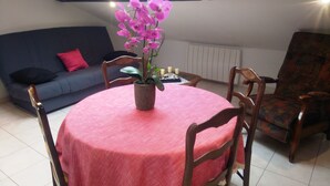 A dining area with a round table covered by a pink tablecloth, surrounded by wooden chairs. A potted orchid plant is placed on the table. The room has a dark blue sofa with red cushions, a patterned armchair, and a white radiator. - 4-person apartment (Docelles)