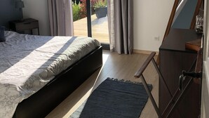 A bedroom with a bed, a desk, a chair, a dresser, and a view of a garden and mountains through a sliding door.