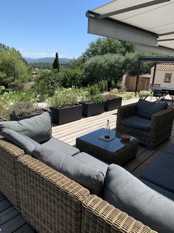 Une terrasse dotée de meubles en osier et de jardinières, offrant une vue sur un jardin et des collines lointaines.