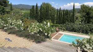 A pool area with a wooden deck, surrounded by lush greenery and tall trees, overlooking a scenic landscape with mountains in the distance.