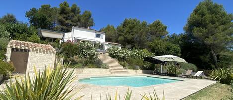 A pool area with a stone deck, a small stone structure, and a white building with a tiled roof in the background.