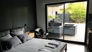 Ein modernes Schlafzimmer mit einem großen Bett, einem Nachttisch, einem Sessel und einer Glastür, die zu einem Außenbereich mit einer Couch und einem Tisch führt.