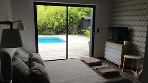 Ein Schlafzimmer mit einer großen Glasschiebetür, die auf eine Holzterrasse und einen Swimmingpool führt. Es gibt ein Bett mit Kissen und einen Fernseher auf einer Holzkommode.