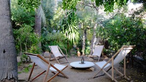 An outdoor seating area with four white lounge chairs and a round table, surrounded by lush greenery and trees.