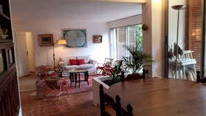 A spacious living room with a white sofa, a red armchair, a coffee table, and a large window with shutters.