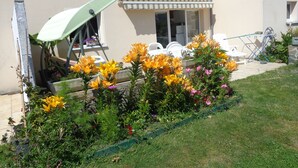 Ein Garten mit gelben und rosafarbenen Blumen, eine Terrasse mit weißen Stühlen und ein Gebäude mit beiger Fassade und grünen Fensterläden.