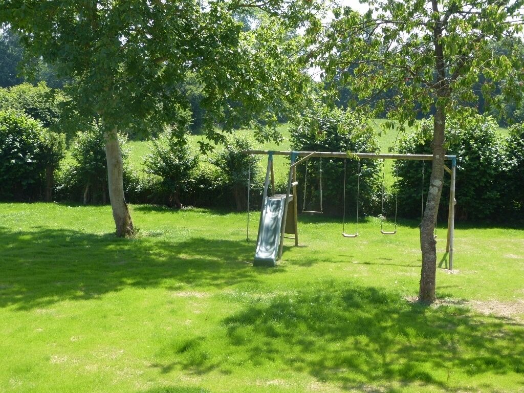 Een speelplaats met een glijbaan en schommels in een grasveld, omringd door bomen.