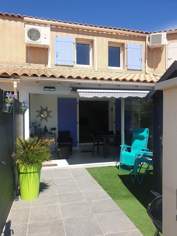A covered patio with outdoor furniture, a small lawn, and a building with a tiled roof and blue shutters.
