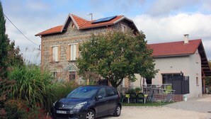 Una casa de piedra con techo de tejas rojas, un coche aparcado y un jardín con diversas plantas y flores.