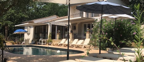 Ein Poolbereich mit Liegestühlen und Sonnenschirmen, umgeben von Grünflächen und einem Haus mit Balkon.