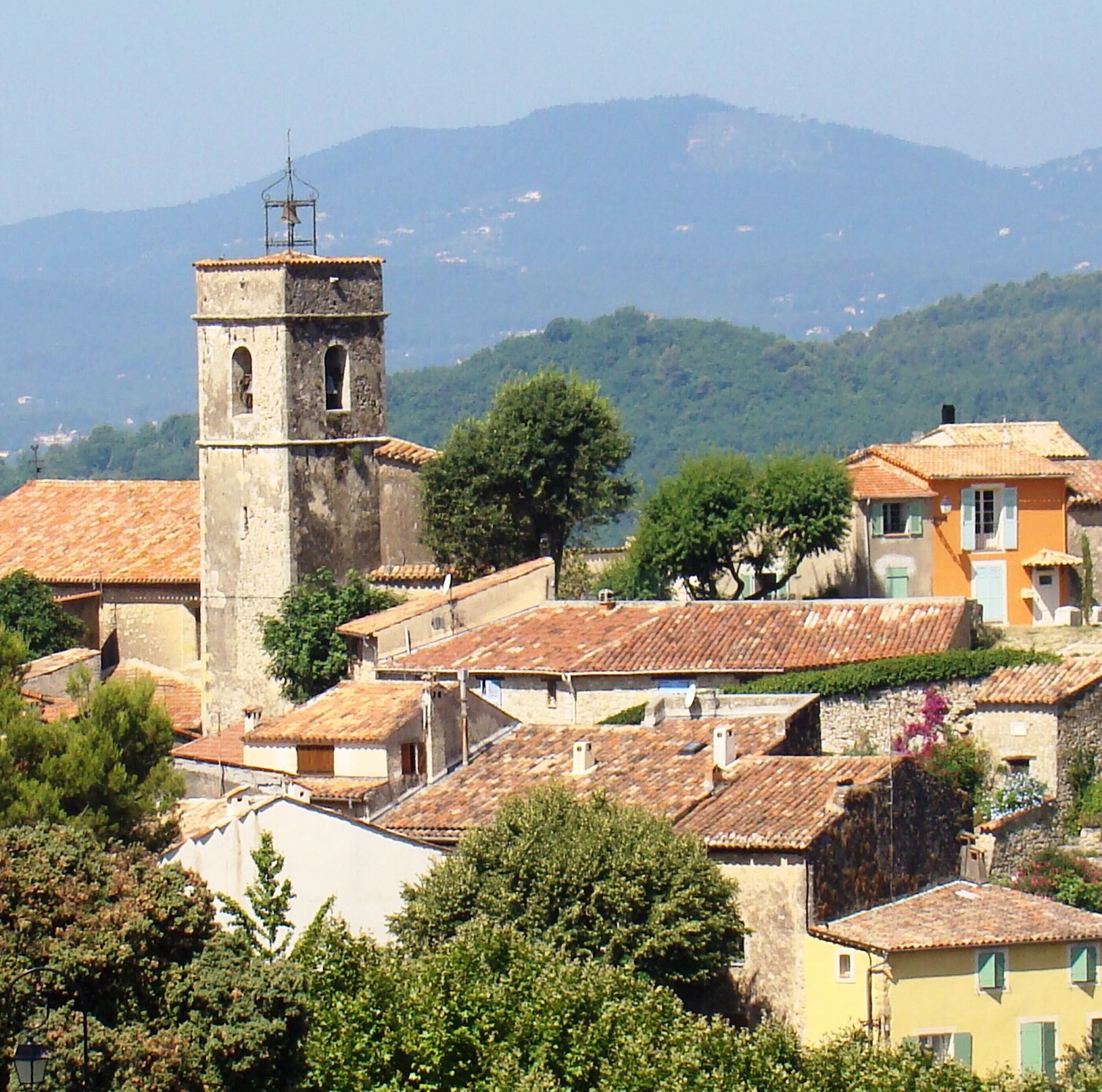 A historic town with a prominent bell tower, traditional buildings with terracotta roofs, and lush greenery.