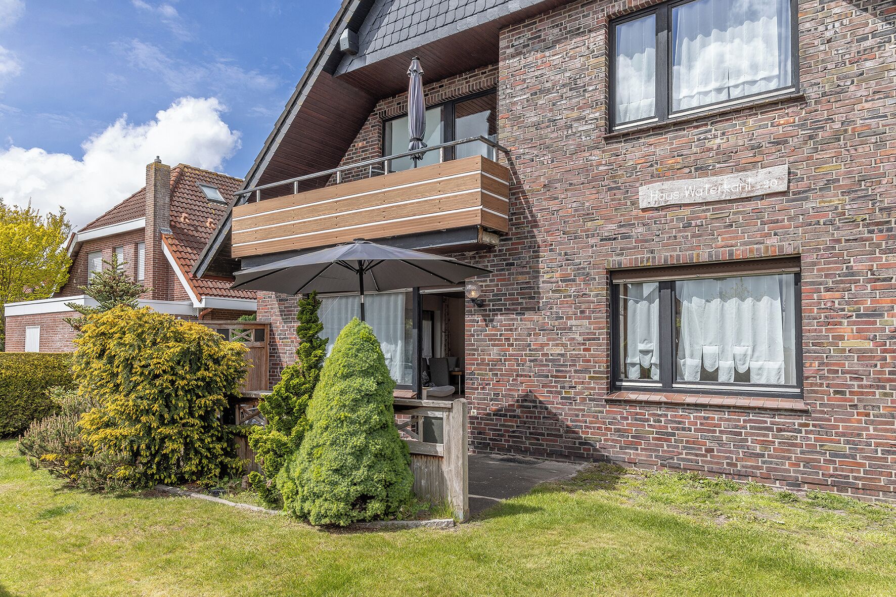 A two-story brick building with a balcony, a sign reading 'Haus Wasserkant,' and a well-maintained garden.
