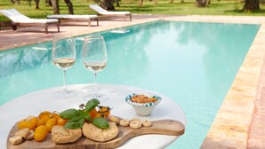 A poolside setting with two glasses of white wine, a bowl of snacks, and a wooden board with various foods.