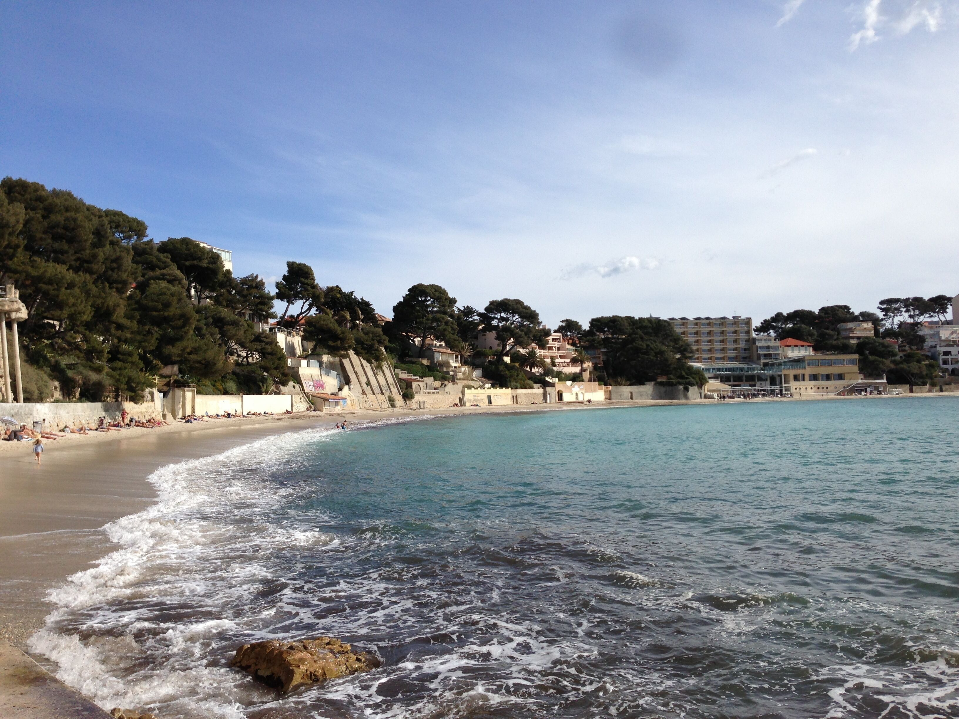 Una playa costera con aguas transparentes, una orilla arenosa y edificios al fondo.