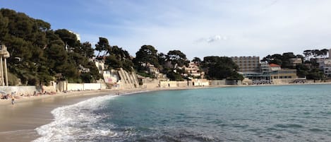 Una playa costera con aguas transparentes, una orilla arenosa y edificios al fondo.